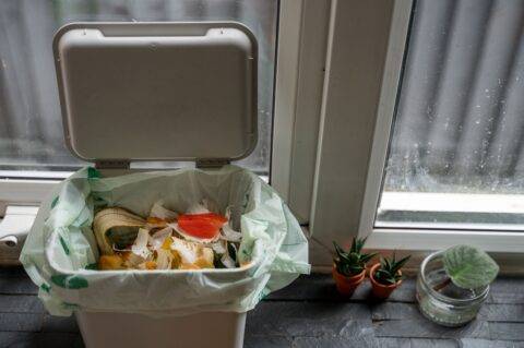 An open garbage bing with some food garbage at the top next to a window.