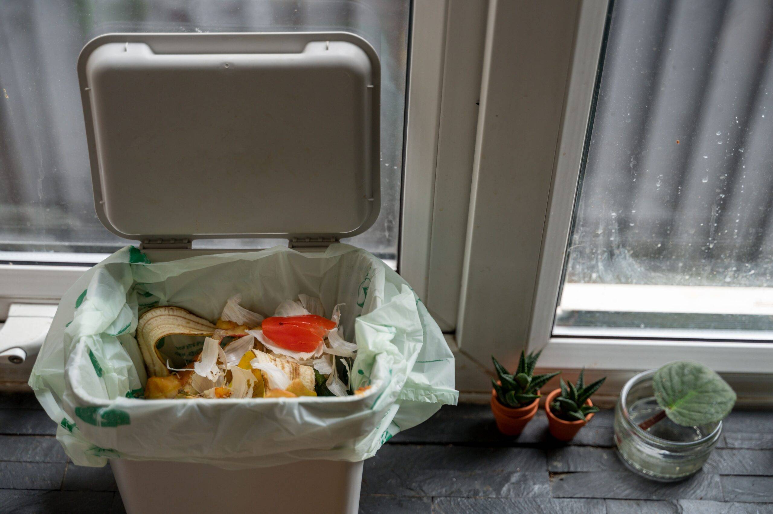 An open garbage bing with some food garbage at the top next to a window.