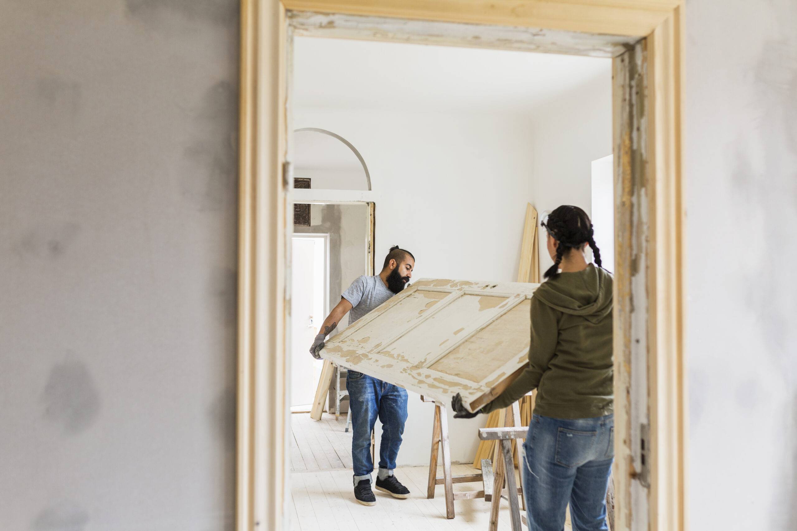 A photo shot through a doorway of a couple doing home renovations carrying either end of a door through an empty room.