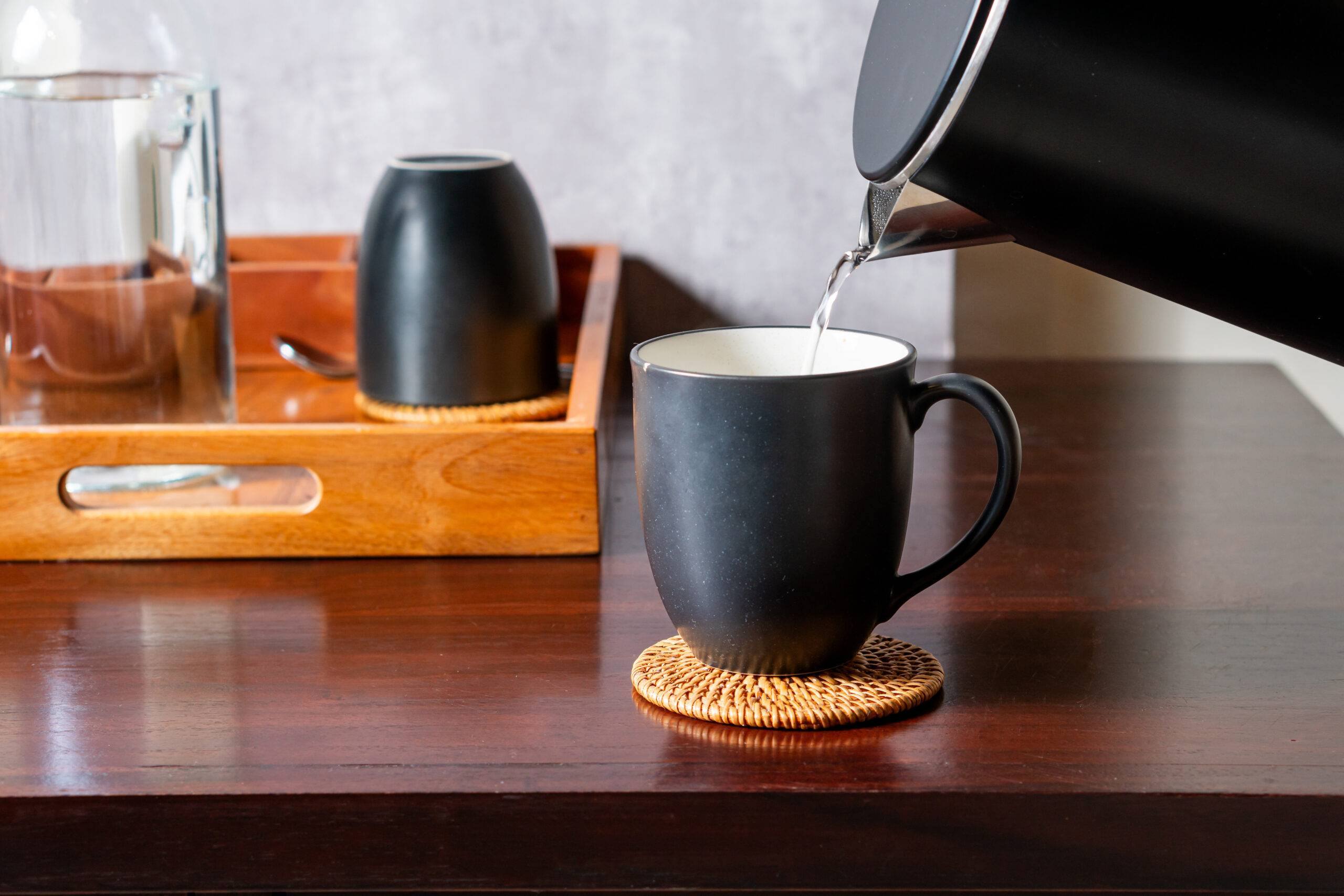A black coffee mug placed on a woven coaster, with hot water being poured from a kettle. In the background, there is a wooden tray with a glass and another black cup. The setting is cozy