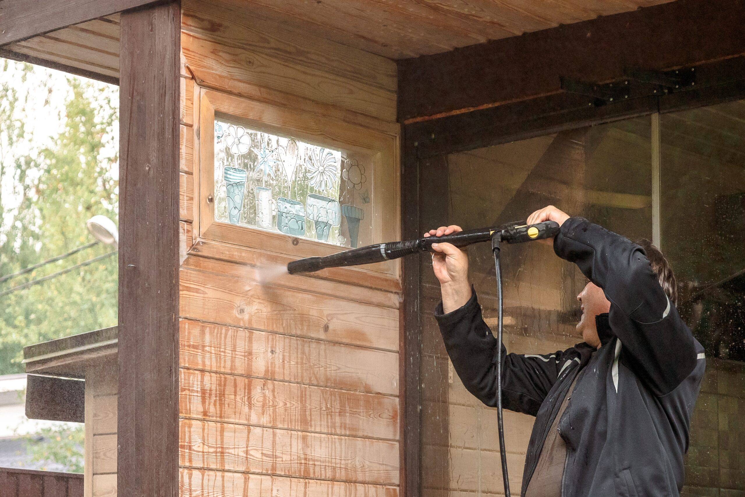 A person raising a pressure washer to wash a wooden outdoor wall.