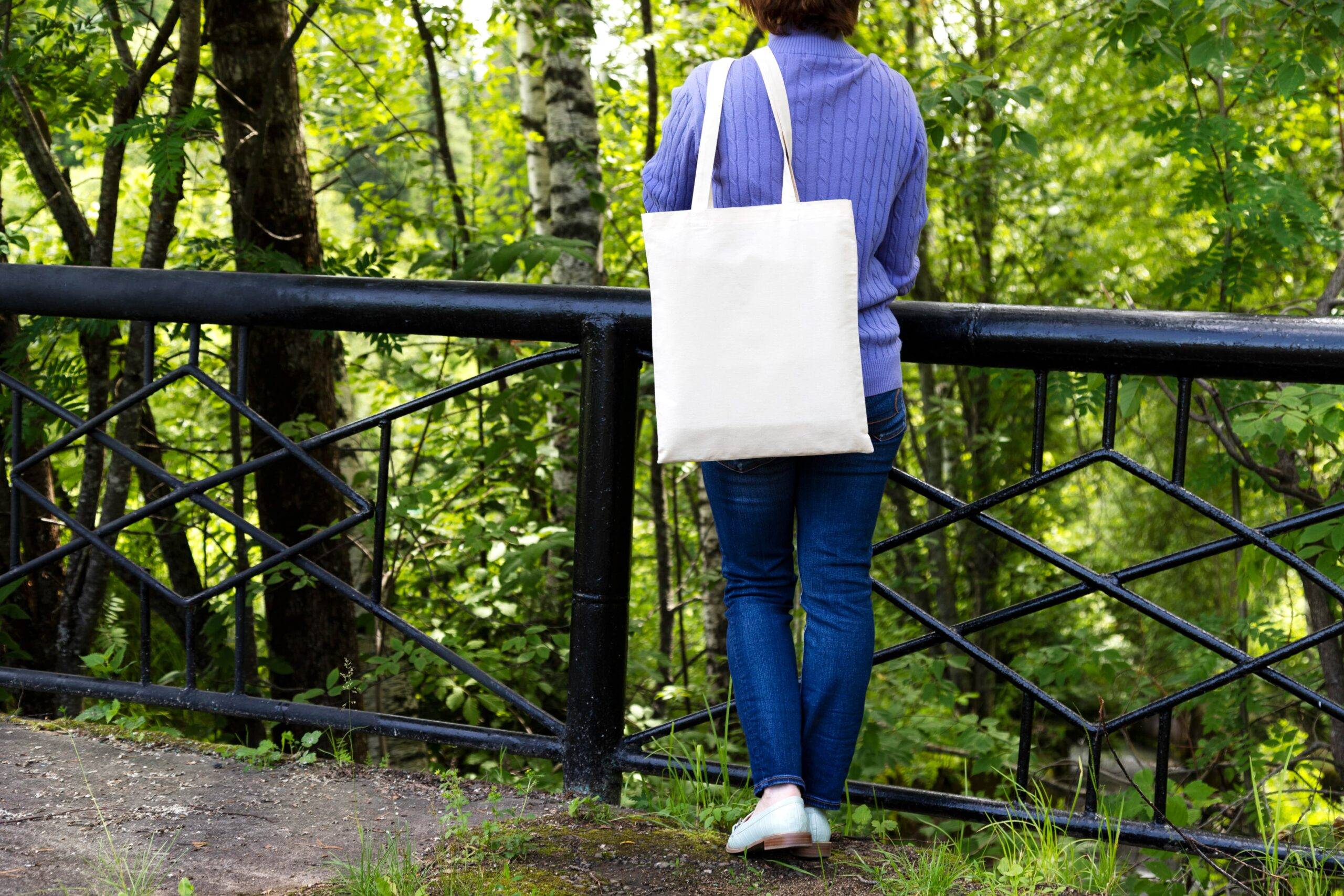 A woman standing outside in a park, leaning against a railing, back to the camera, white tote bag strung over her shoulder.