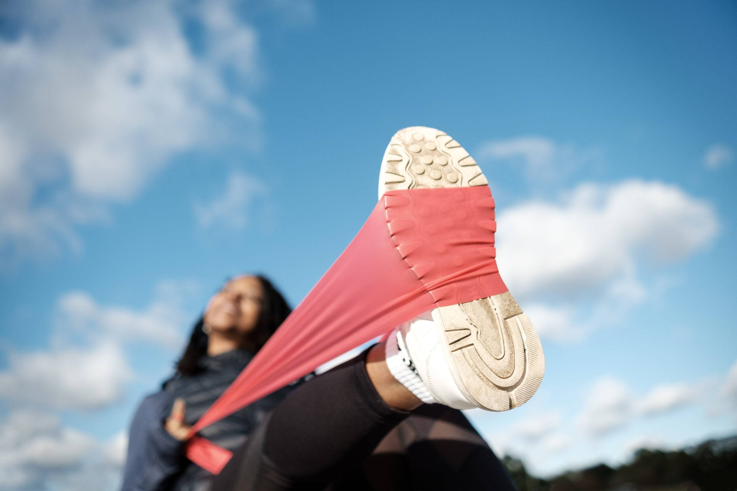 Black woman is using a resistance band outdoors. Sunny autumn day. We have a close-up view of the right foot with the resistance band.