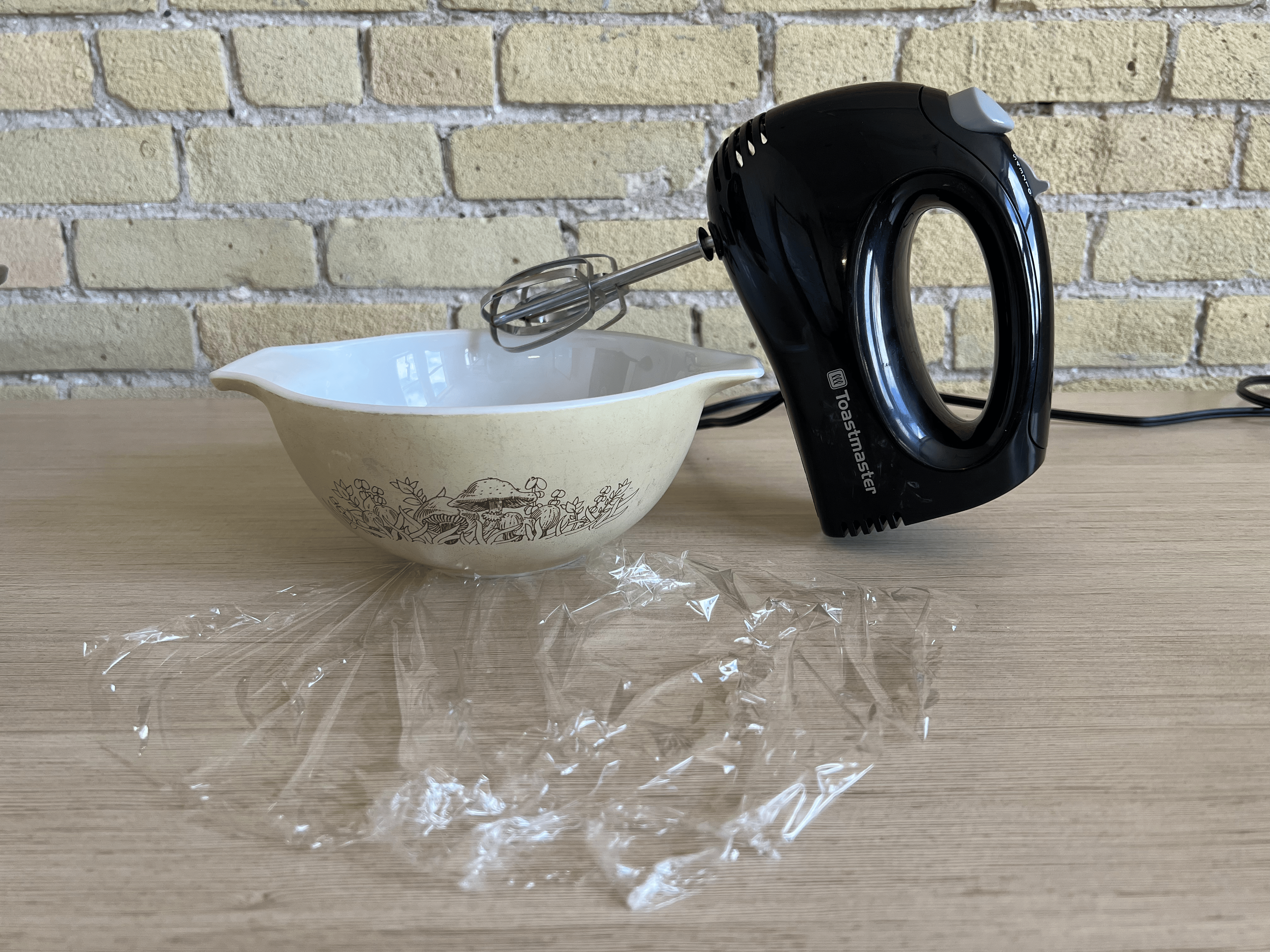 A bowl, a hand mixer, and a piece of plastic wrap all on a table.