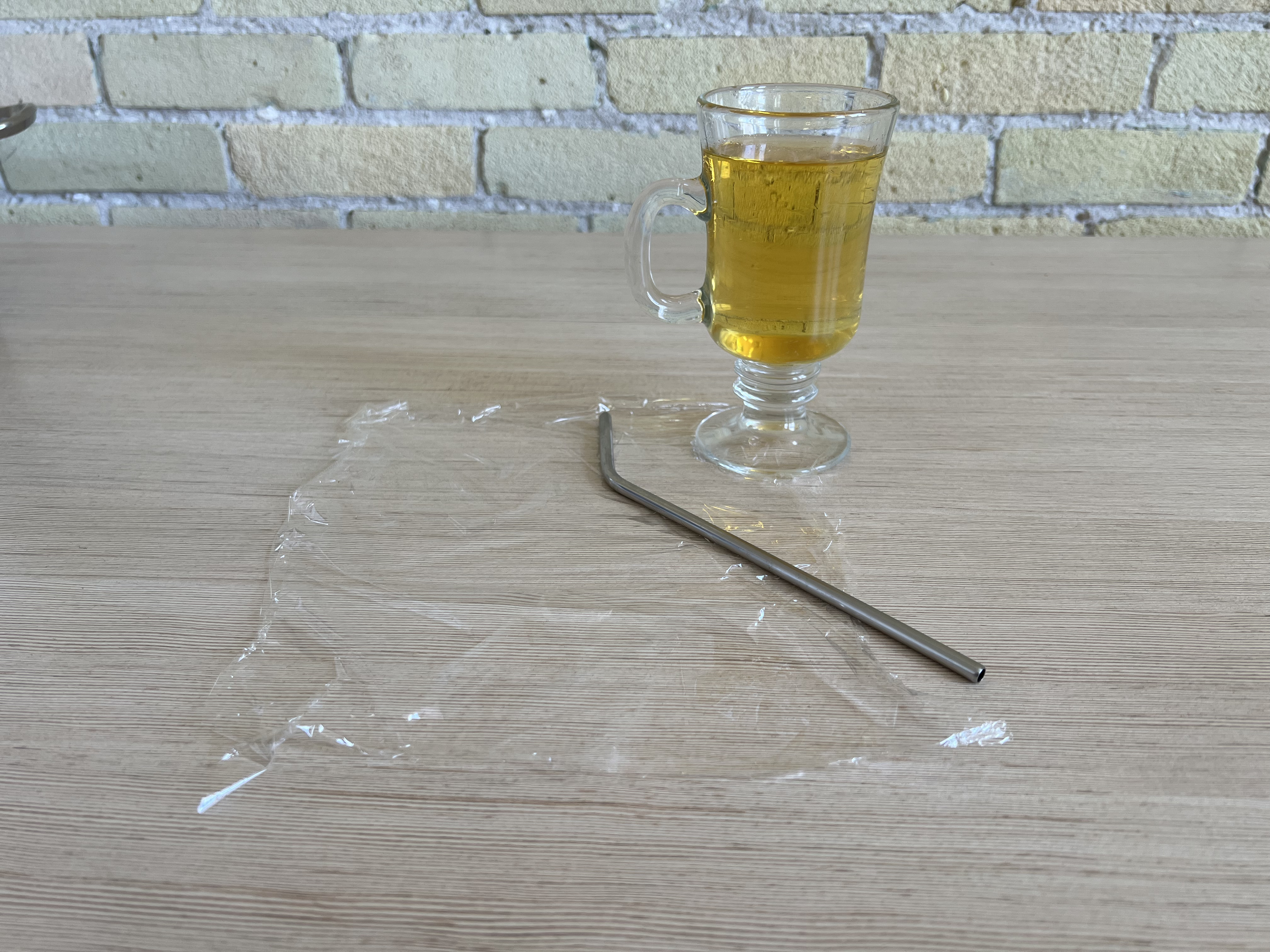 A cup of apple juice, a metal straw, and a piece of plastic wrap on a table.