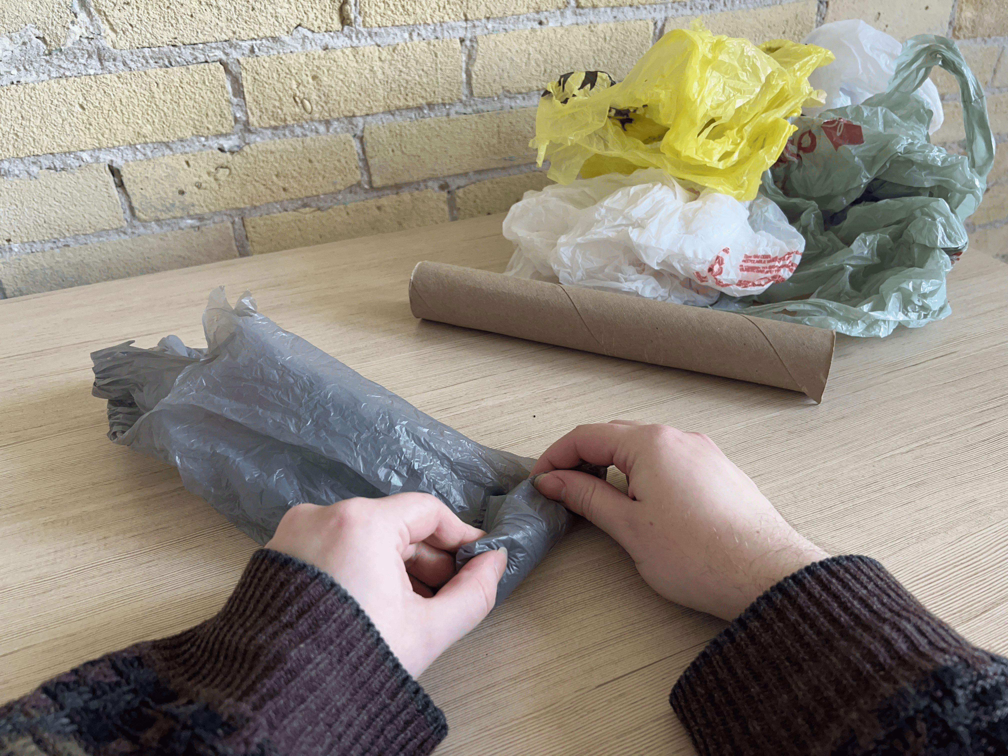 A pair of hands rolling up a plastic bag, a paper towel roll and small pile of other plastic bags visible as well.