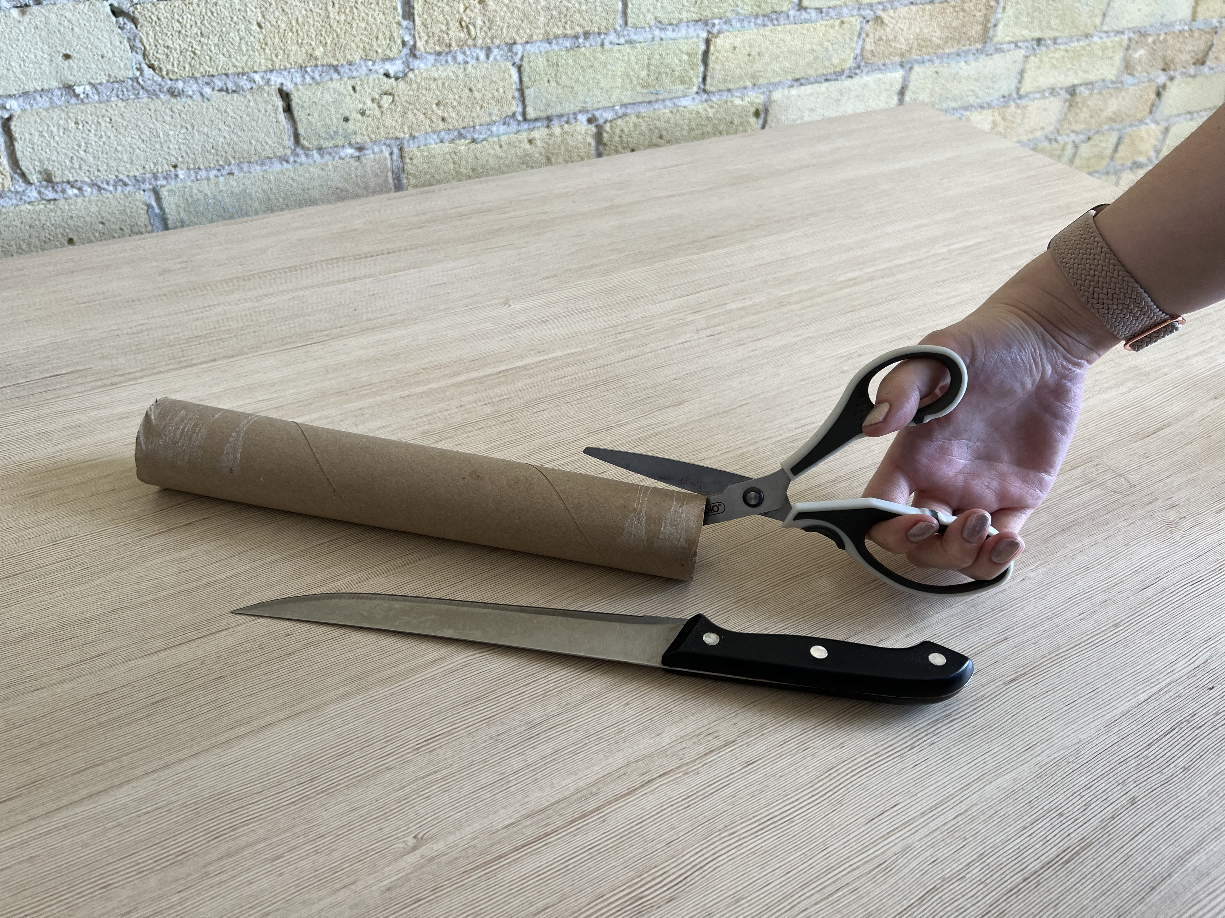 A hand cutting a paper towel roll that's sitting next to a cooking knife.