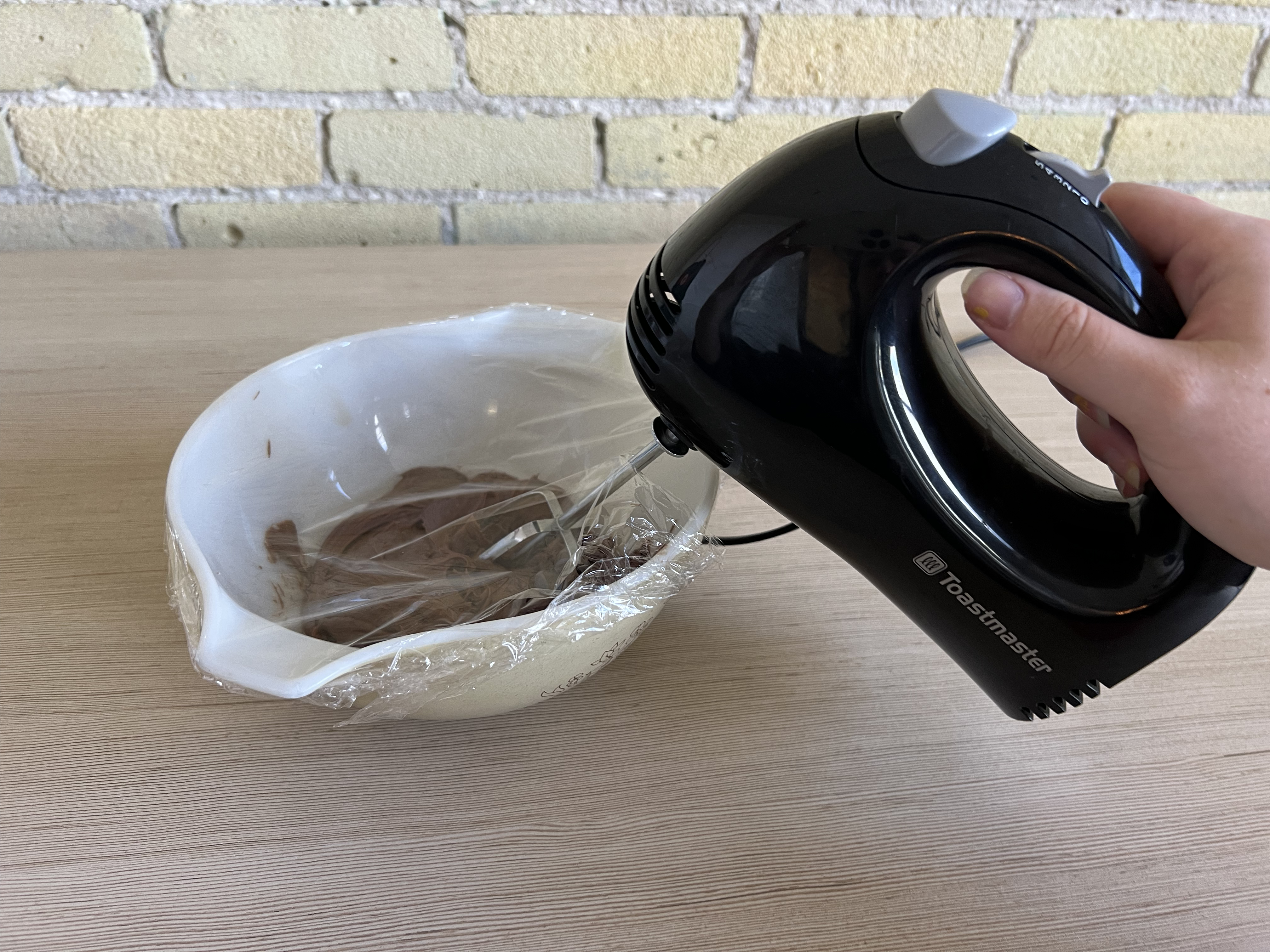 A hand mixer in a bowl of icing with plastic wrap covering the top.