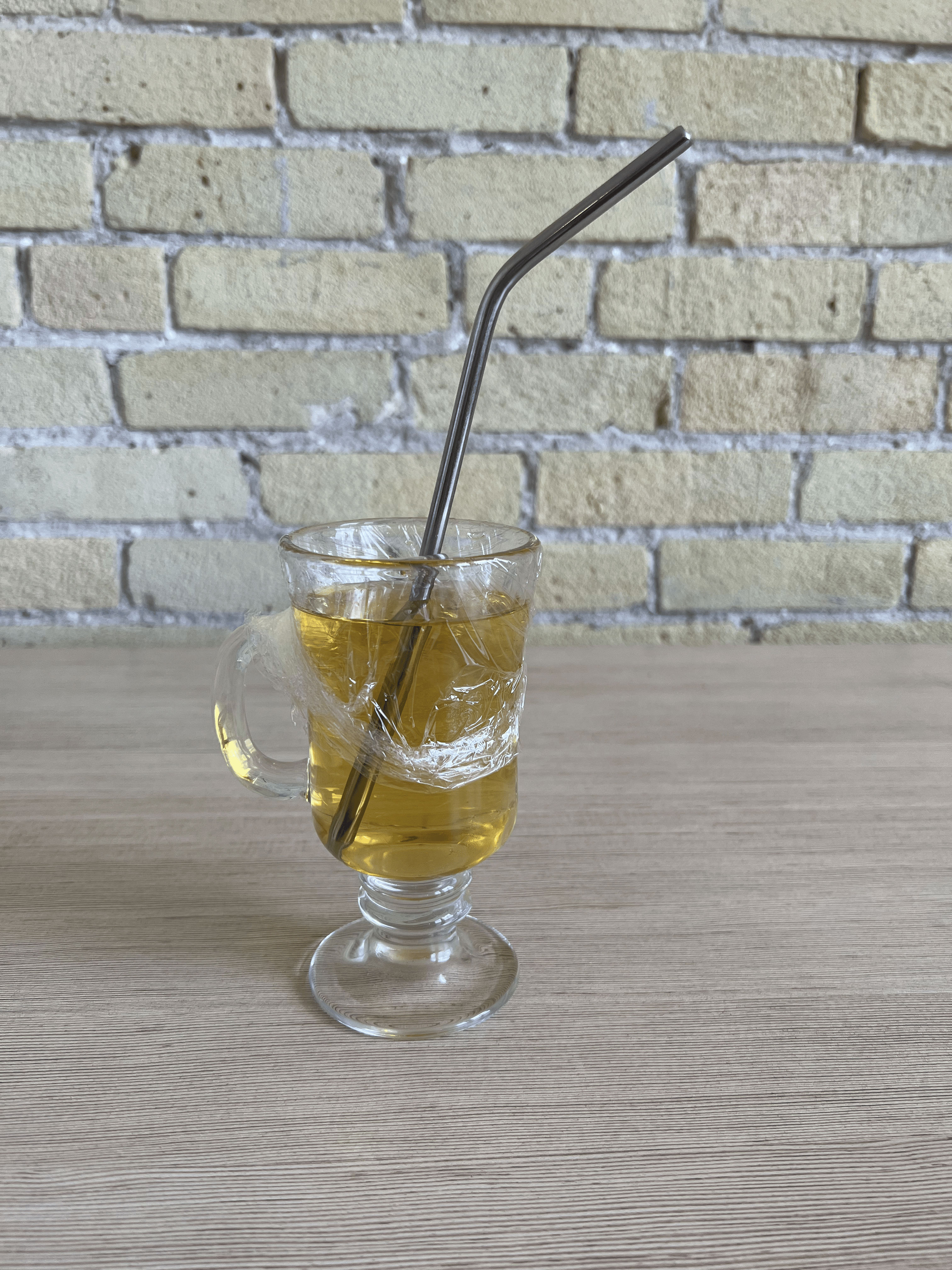 A glass filled with apple juice, a piece of plastic wrap covering the top, a straw stabbed through it.