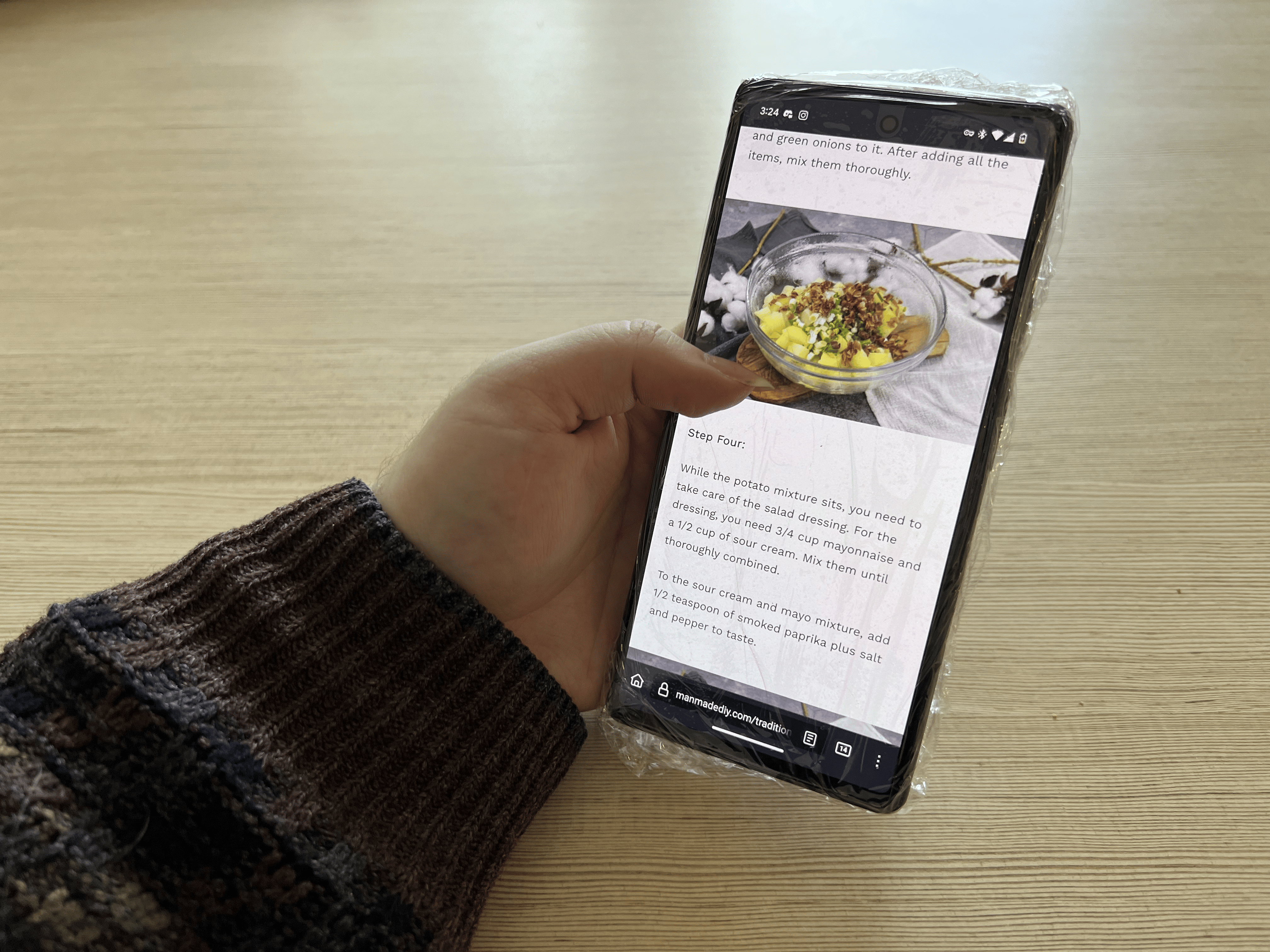 A hand holding the phone wrapped in plastic wrap, scrolling through a recipe.